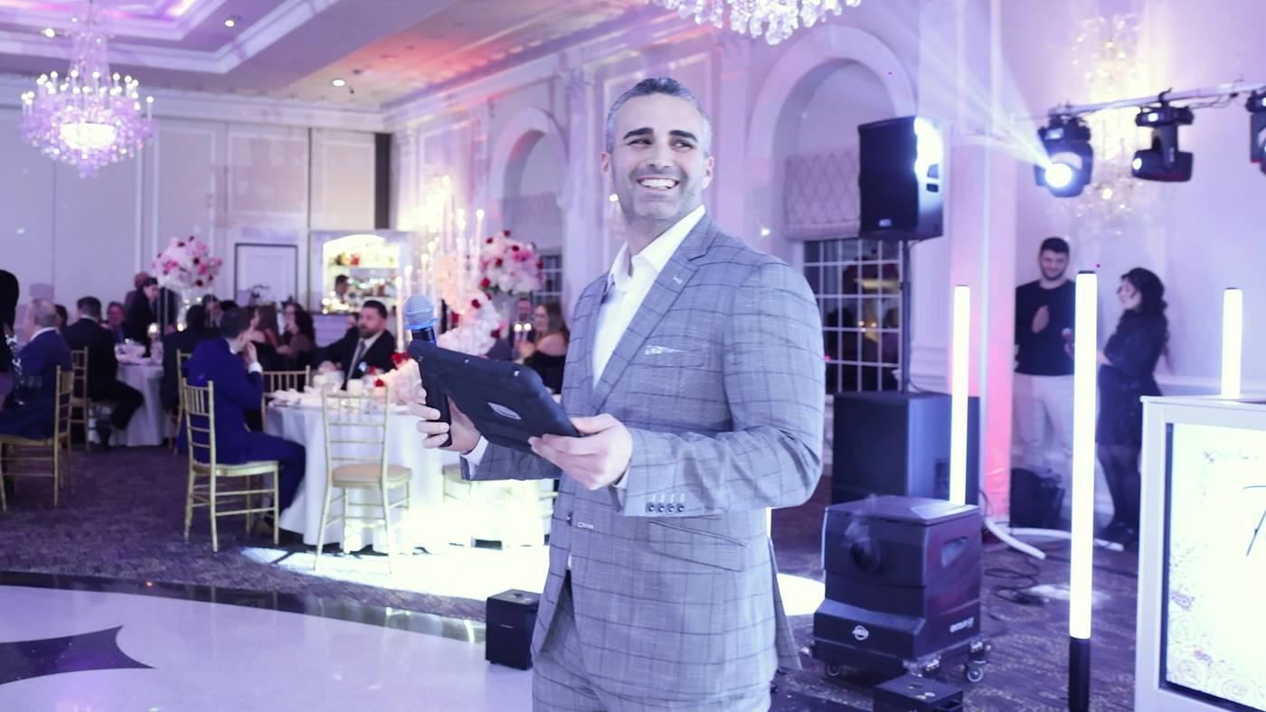 Joey Disaronno on the microphone as MC in a chandeliered ballroom, with the DJ booth, speakers and pink uplighting set behind him